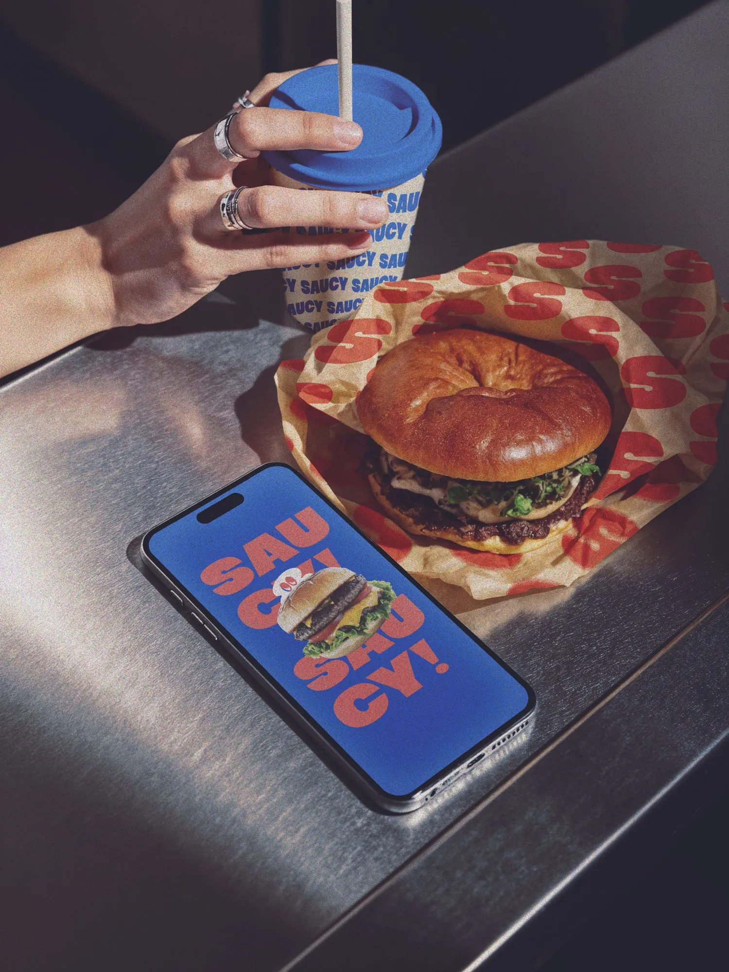 Combo de Saucy sobre un mostrador de acero: vaso, hamburguesa envuelta y la app en el celular
