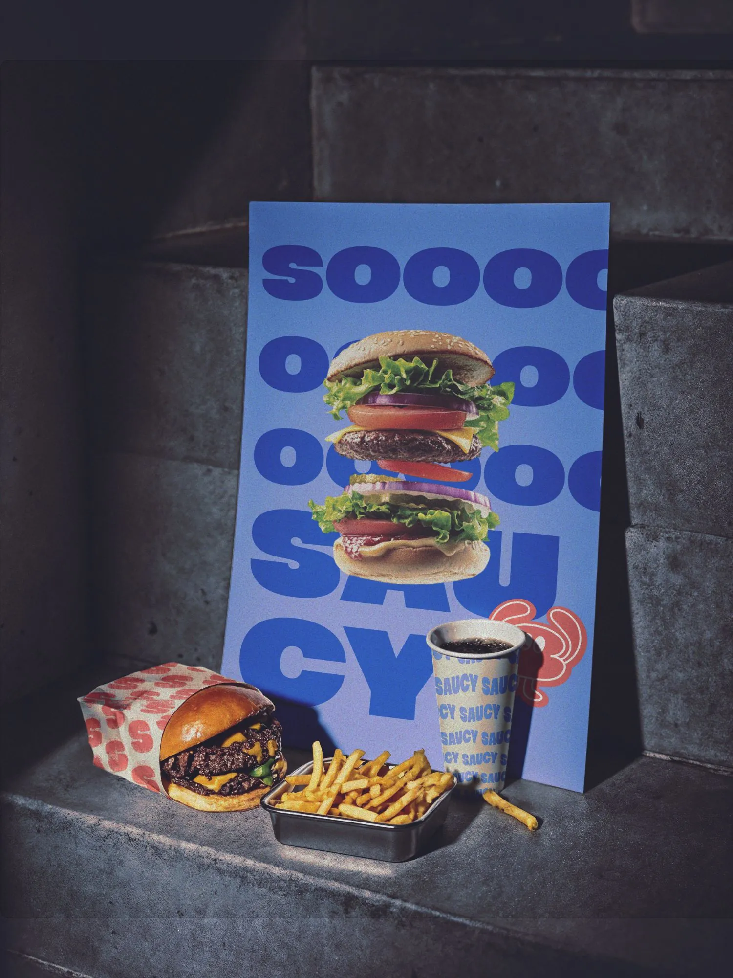 Afiche SOOOO SAUCY! apoyado junto a la hamburguesa, las papas y el vaso de la marca