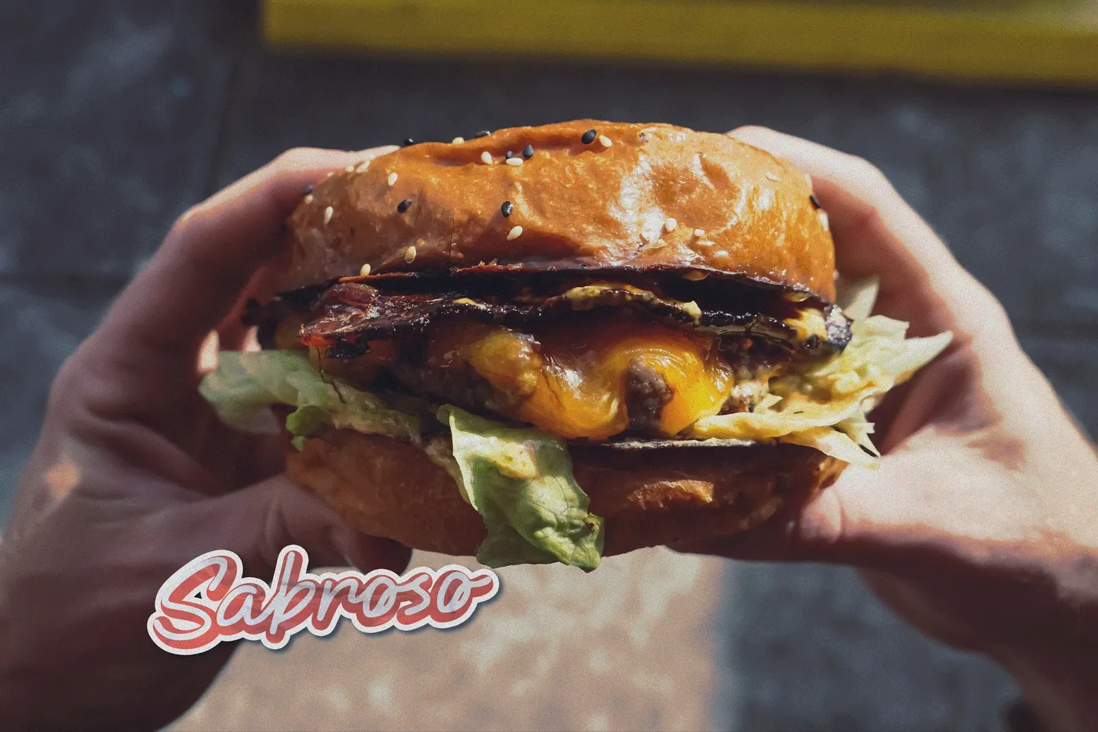 Hamburguesa sostenida en las manos con el sticker “Sabroso” de Saucy
